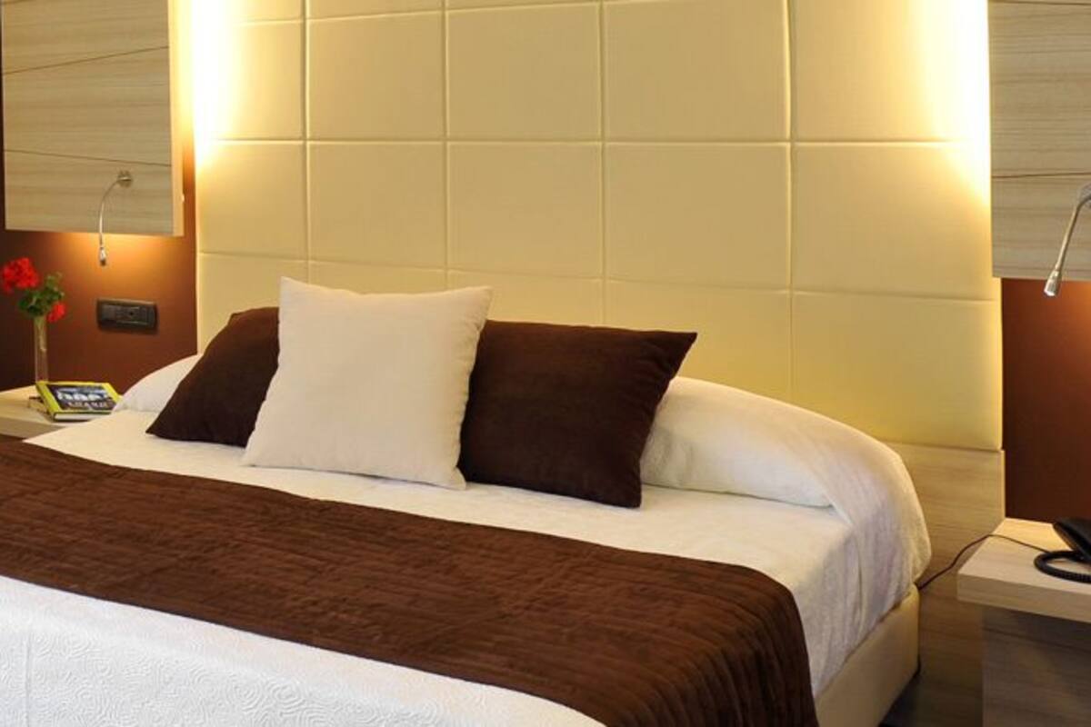 Una habitación de hotel con una cama, una silla, una mesita de noche con lámpara y otro mesita con un teléfono.