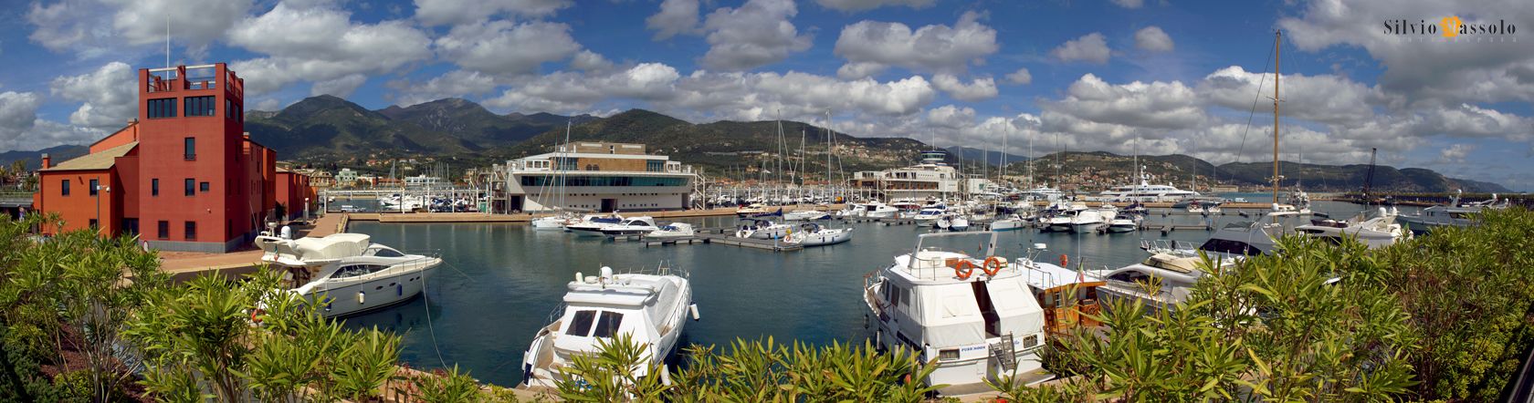 A marina with multiple boats docked, a red building, and a mountainous backdrop.