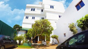 A white building with multiple windows and a green roof. There are two cars parked in front, and a tree in the center. - Colombo Residency (Colombo)