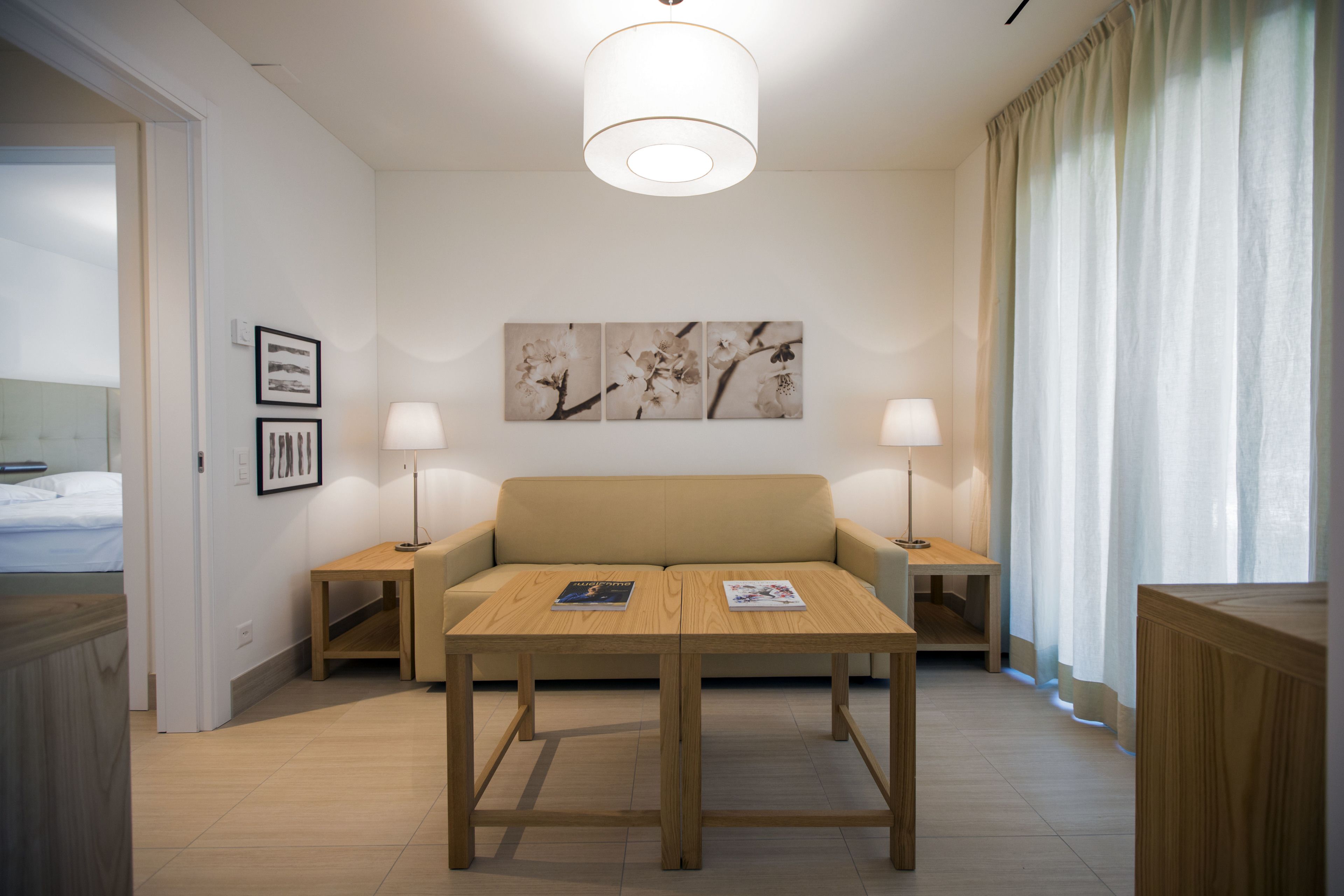 Ein modernes Hotelzimmer mit einem beigefarbenen Sofa, einem Holztisch, zwei Nachttischlampen und einem großen Fenster mit transparenten Vorhängen.