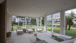 A modern, open-plan living area with large windows, wicker furniture, and a view of the surrounding landscape. - Delta Resort Apartments (Ascona)