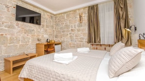 A bedroom with a stone wall, a flat-screen TV, a wooden desk, a bed with a white duvet, and a window with curtains. - Villa Desideria (Split)