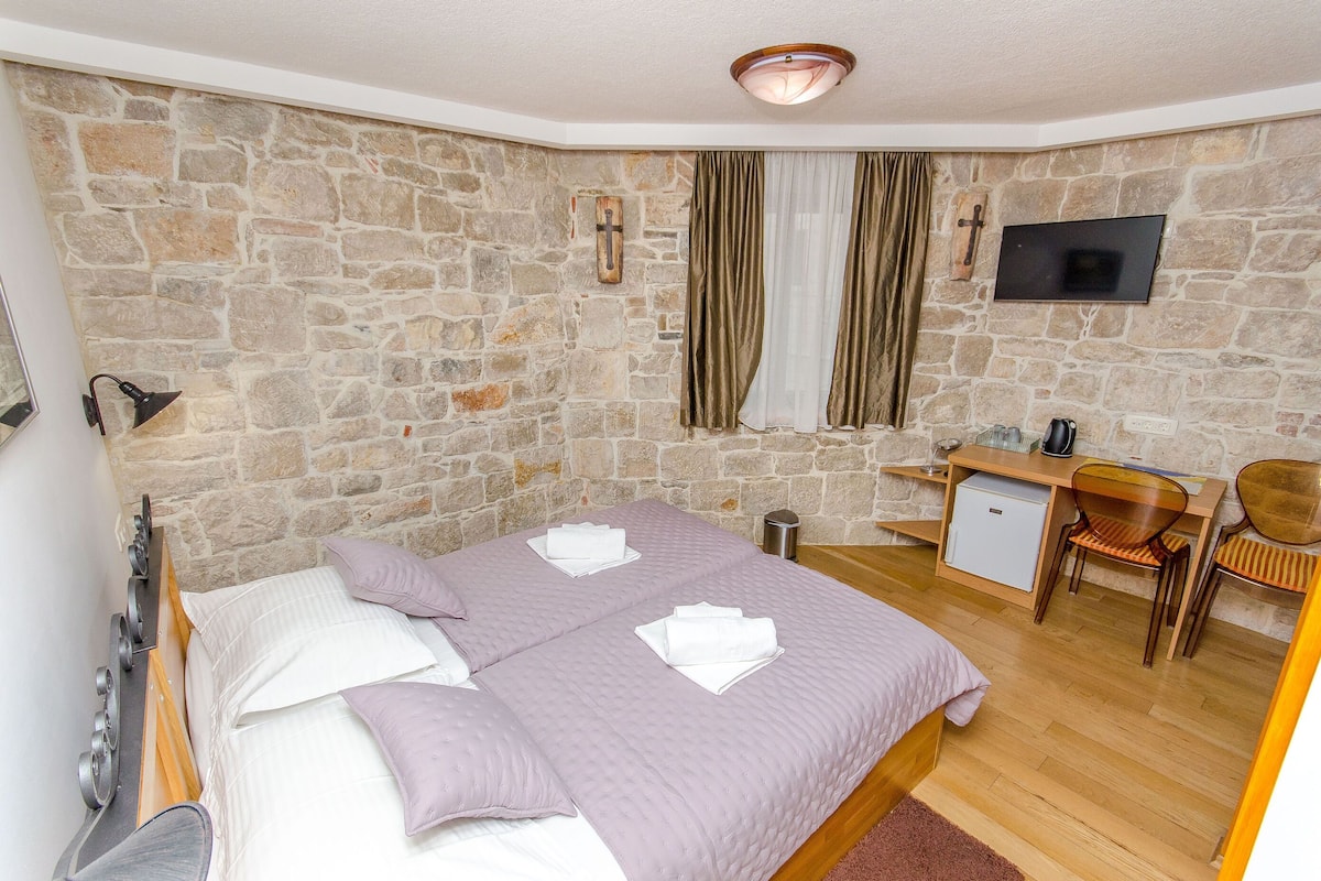 A bedroom with a stone wall, a bed with white linens, a desk with a chair, a television, and a small refrigerator.