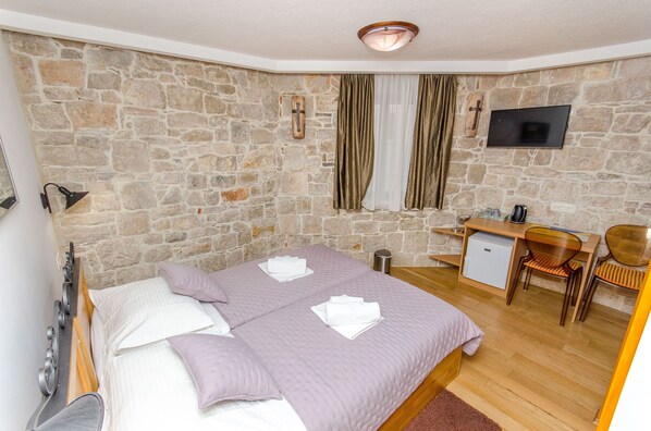 A bedroom with a stone wall, a bed with white linens, a desk with a chair, a television, and a small refrigerator. - Villa Desideria (Split)