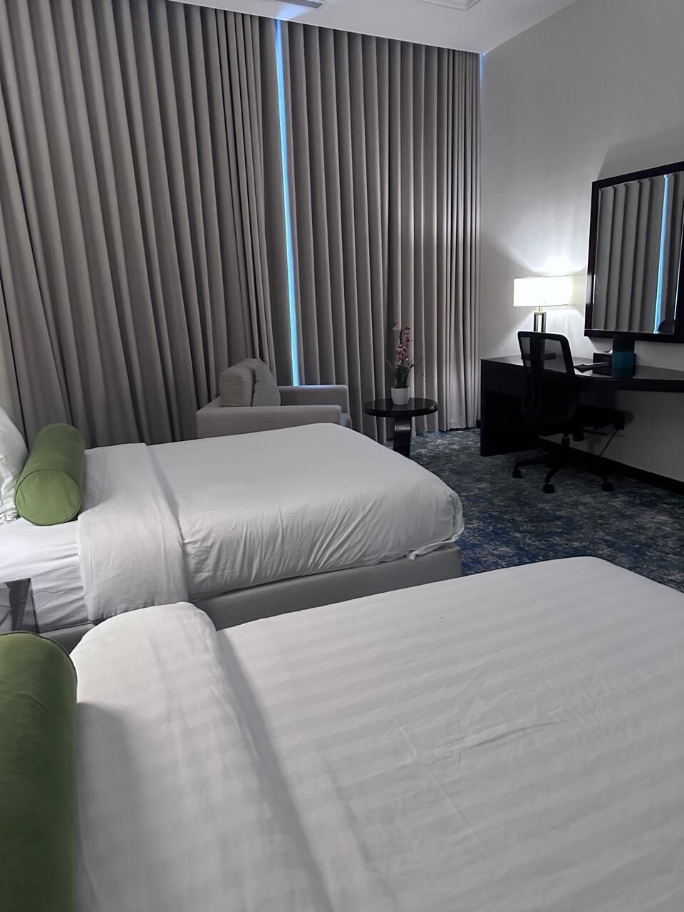 A hotel room with two beds, a desk, a chair, a lamp, and a large window with curtains.