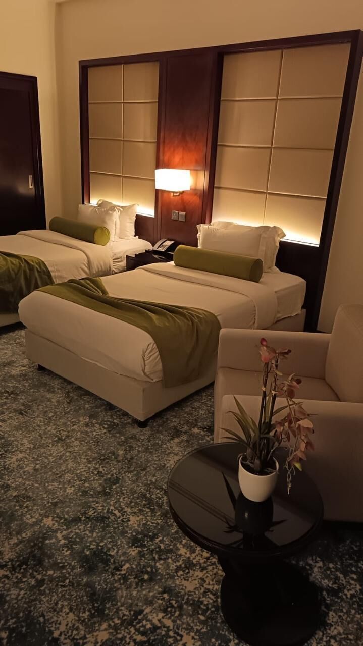 A hotel room with two beds, a sofa, a small table with a plant, and a wall-mounted lamp.