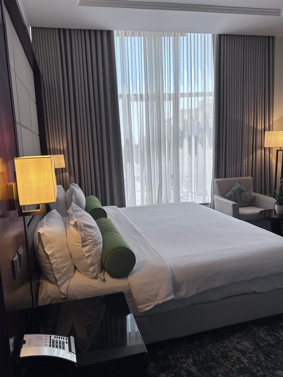 Ein Hotelzimmer mit einem großen Bett, einem Schreibtisch mit Lampe, einem Sessel und Blick durch leichte Vorhänge.