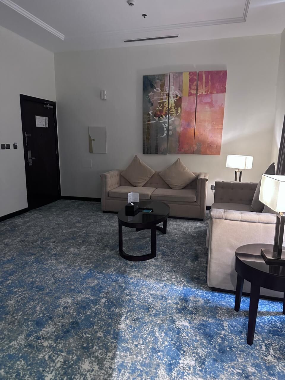 Een hotelkamer met een beige bank, een klein rond tafeltje, een lamp en een schilderij aan de muur.