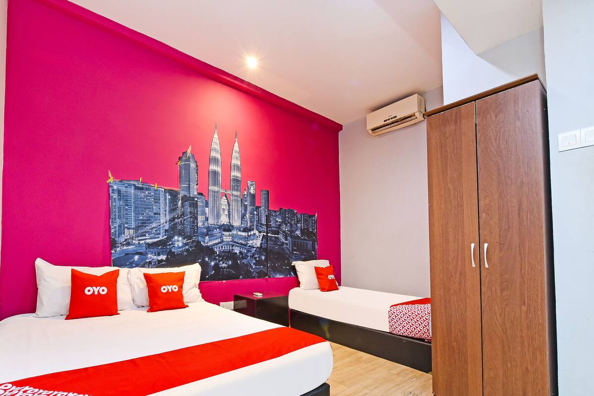 A hotel room with a vibrant pink wall, a large bed with white and red bedding, a wooden wardrobe, and a cityscape painting.