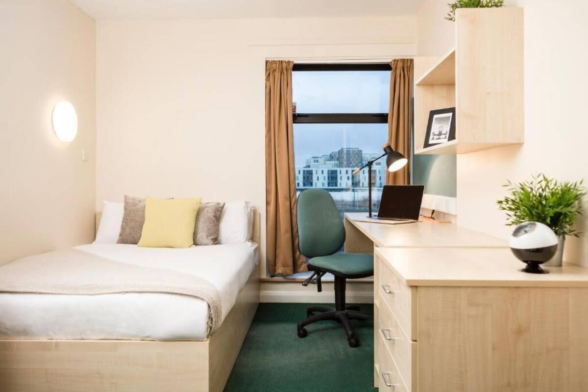 A hotel room with a bed, desk, chair, and a view of the city through the window.