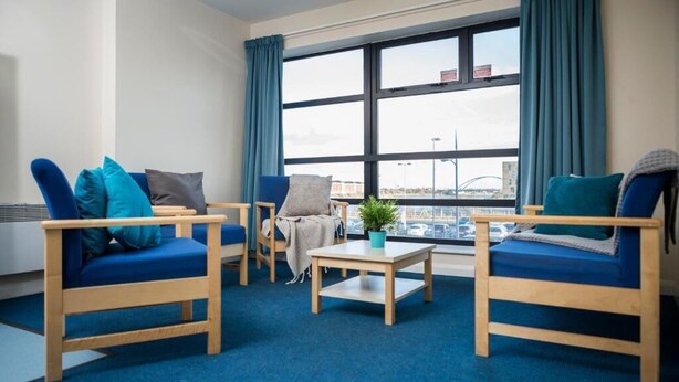 A room with two blue sofas, a wooden coffee table, and a view of a cityscape through large windows with blue curtains. - Bonington Student Village - Campus Accommodation (Loughborough)