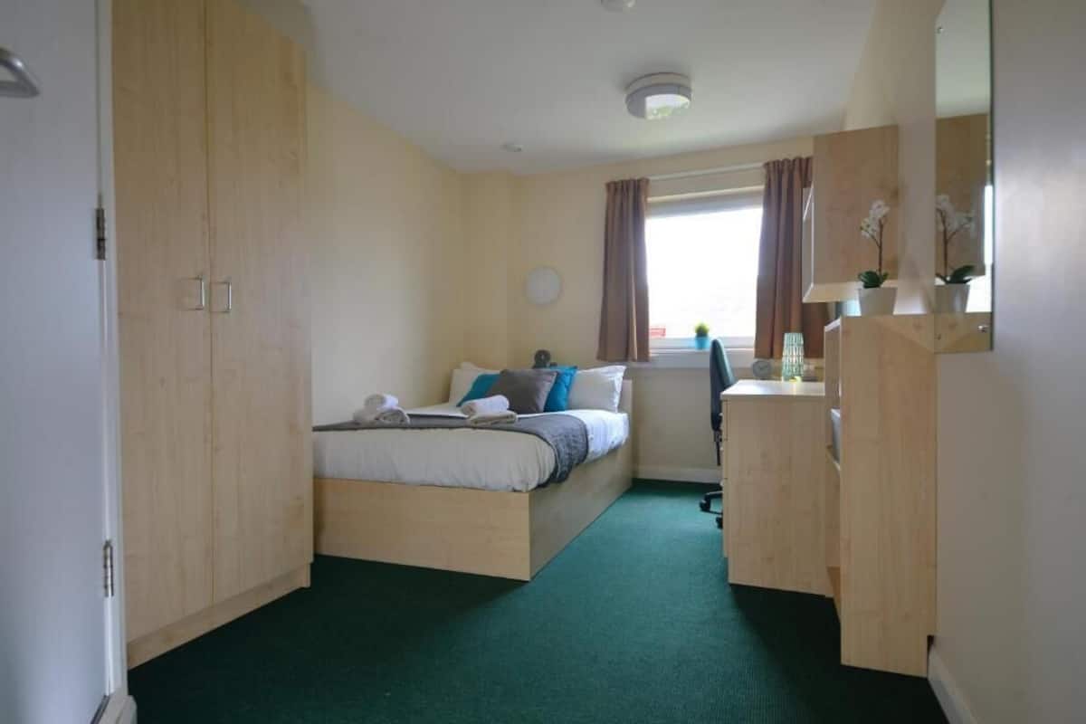 A bedroom with a bed, a wooden wardrobe, a desk, and a window with curtains.