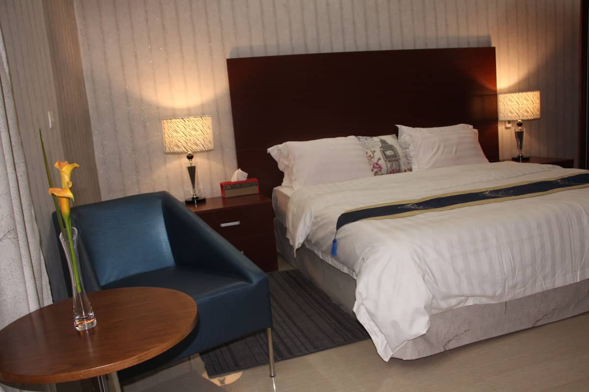 Ein Hotelzimmer mit einem großen Bett, einem Nachttisch mit Lampe, einem kleinen runden Tisch mit einer Vase und einem blauen Sessel.