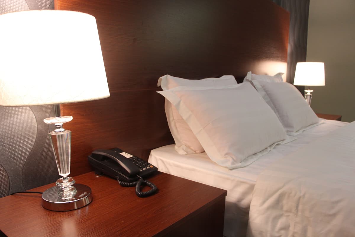 Ein Hotelzimmer mit einem hölzernen Kopfteil, einem Nachttisch mit Lampe und Telefon sowie einem ordentlich bezogenen Bett mit weißen Leinentüchern.