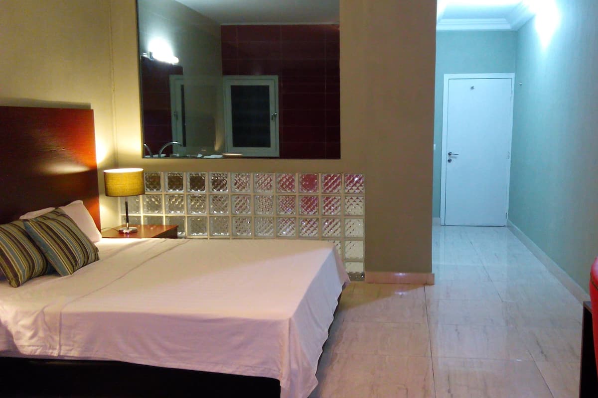 Ein Hotelzimmer mit Bett, Nachttisch mit Lampe, rotem Sessel, Badezimmer mit Spiegel und Fliesen sowie einer Tür zu einem weiteren Raum.