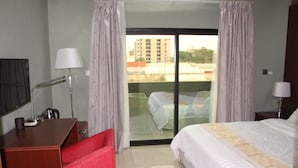 A hotel room with a bed, a desk, a chair, a lamp, and a balcony with a view of buildings. - ELEVEN'S Hôtel & Spa (Abidjan)