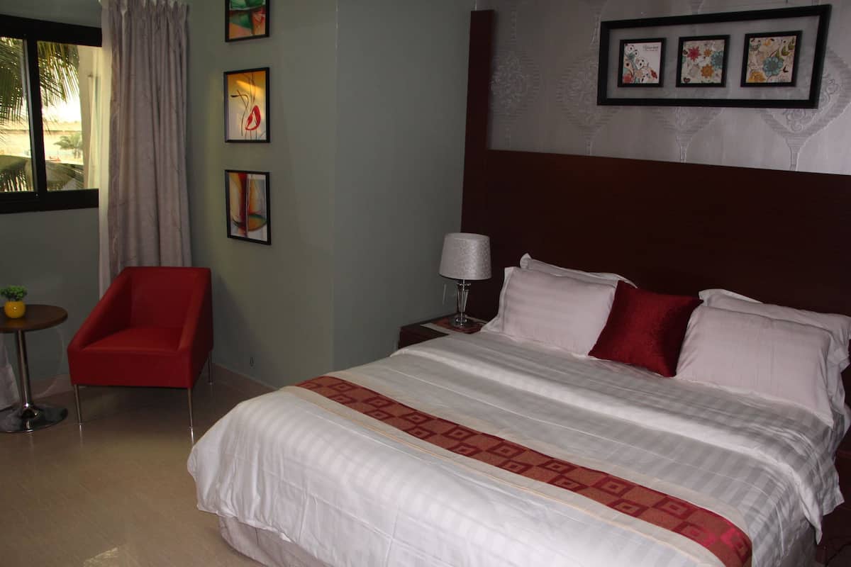 Ein Hotelzimmer mit einem großen Bett, einem roten Sessel, einem Nachttisch mit Lampe sowie gerahmten Kunstwerken an der Wand.