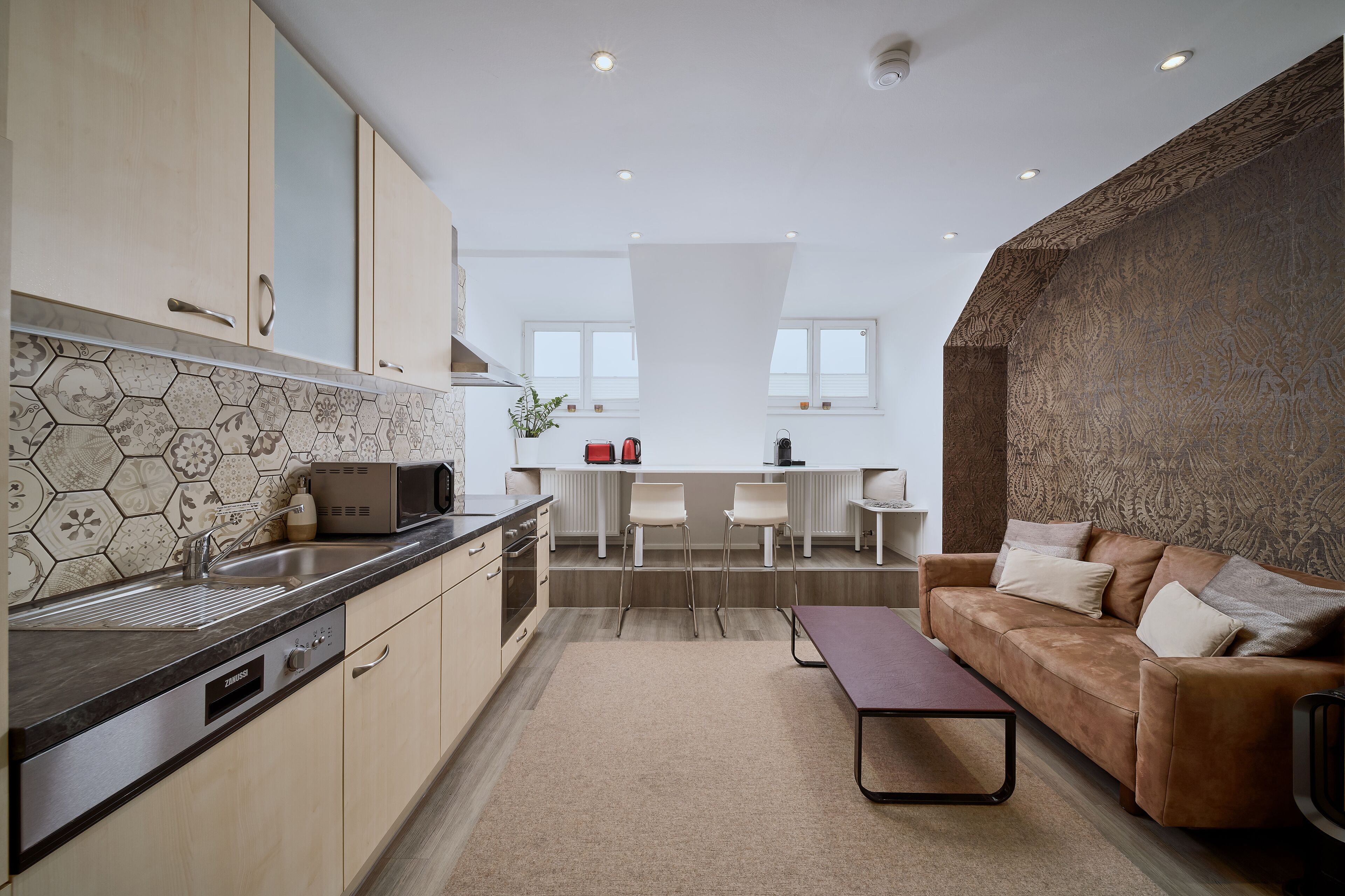 A modern kitchen with a built-in oven, a microwave, and a sink. There is a dining area with a table and chairs. A brown leather sofa is in the living area.