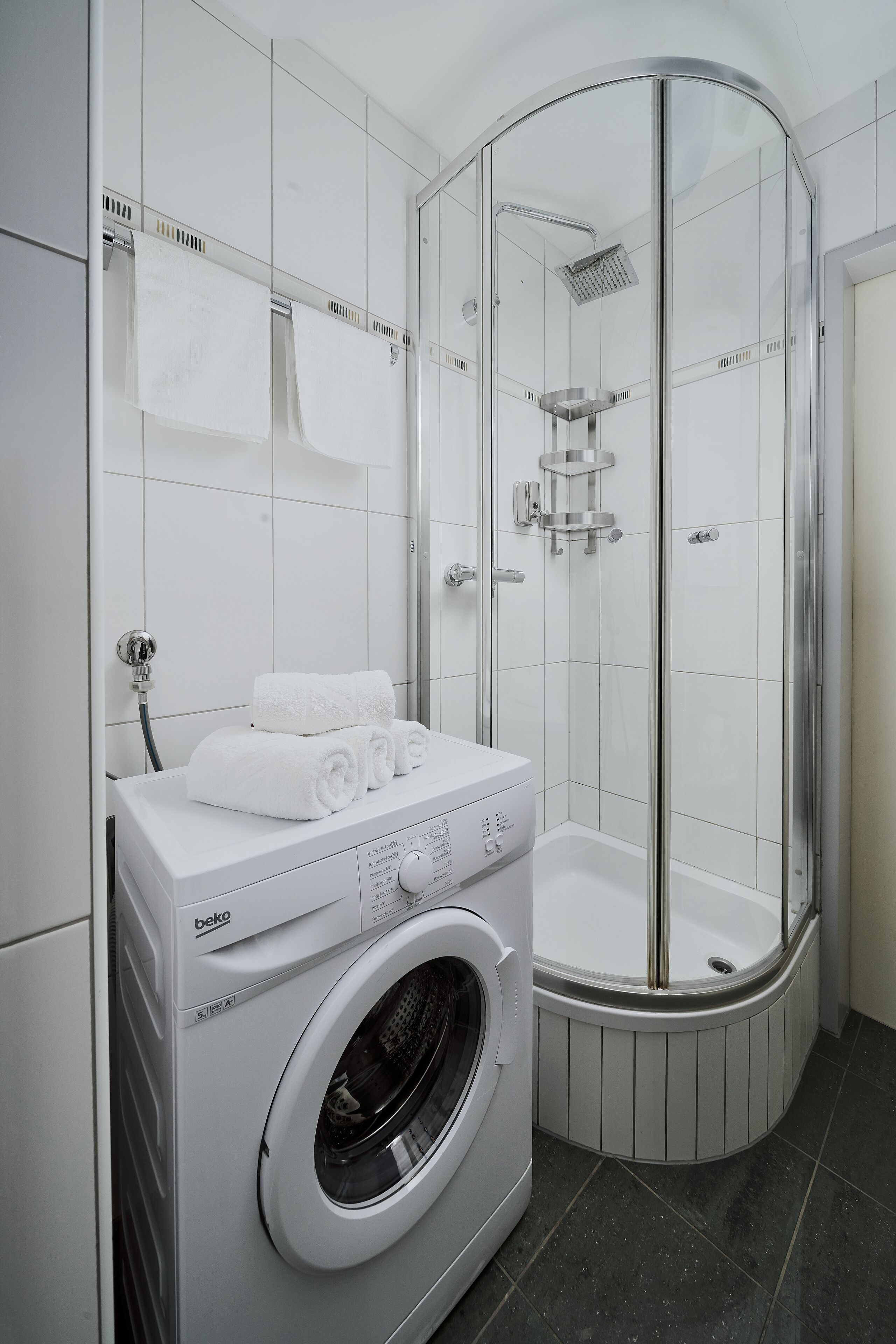 A modern bathroom with a built-in washing machine, a glass-enclosed shower, and a white tiled floor.
