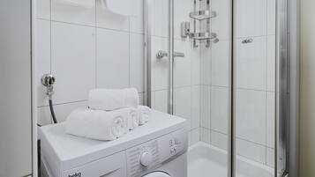 A modern bathroom with a built-in washing machine, a glass-enclosed shower, and a white tiled floor.