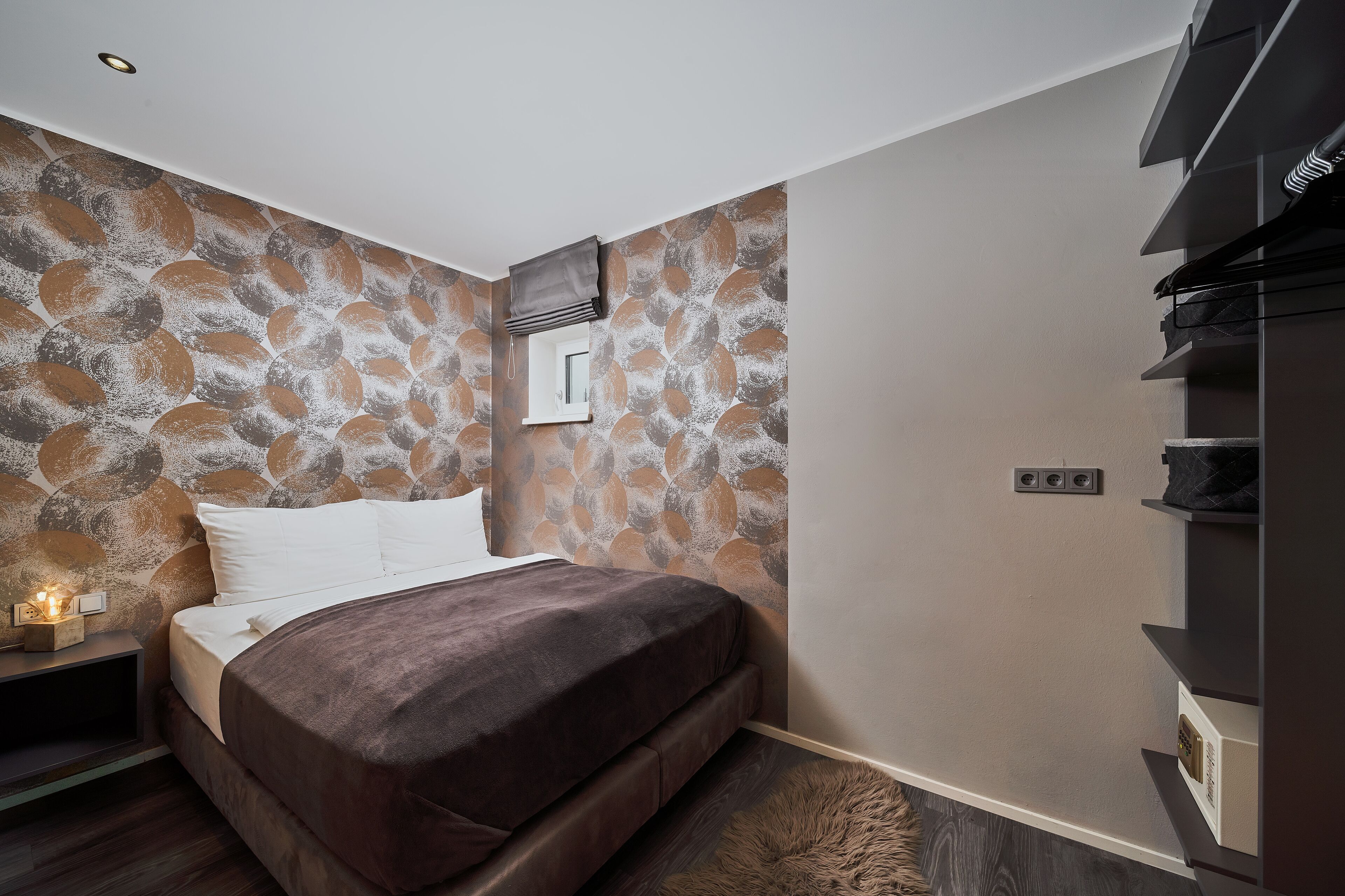 Ein modernes Schlafzimmer mit Wand im Steinoptik, einem Bett mit braunem Kopfteil, einem Nachttisch mit Lampe und einem eingebauten Kleiderschrank.