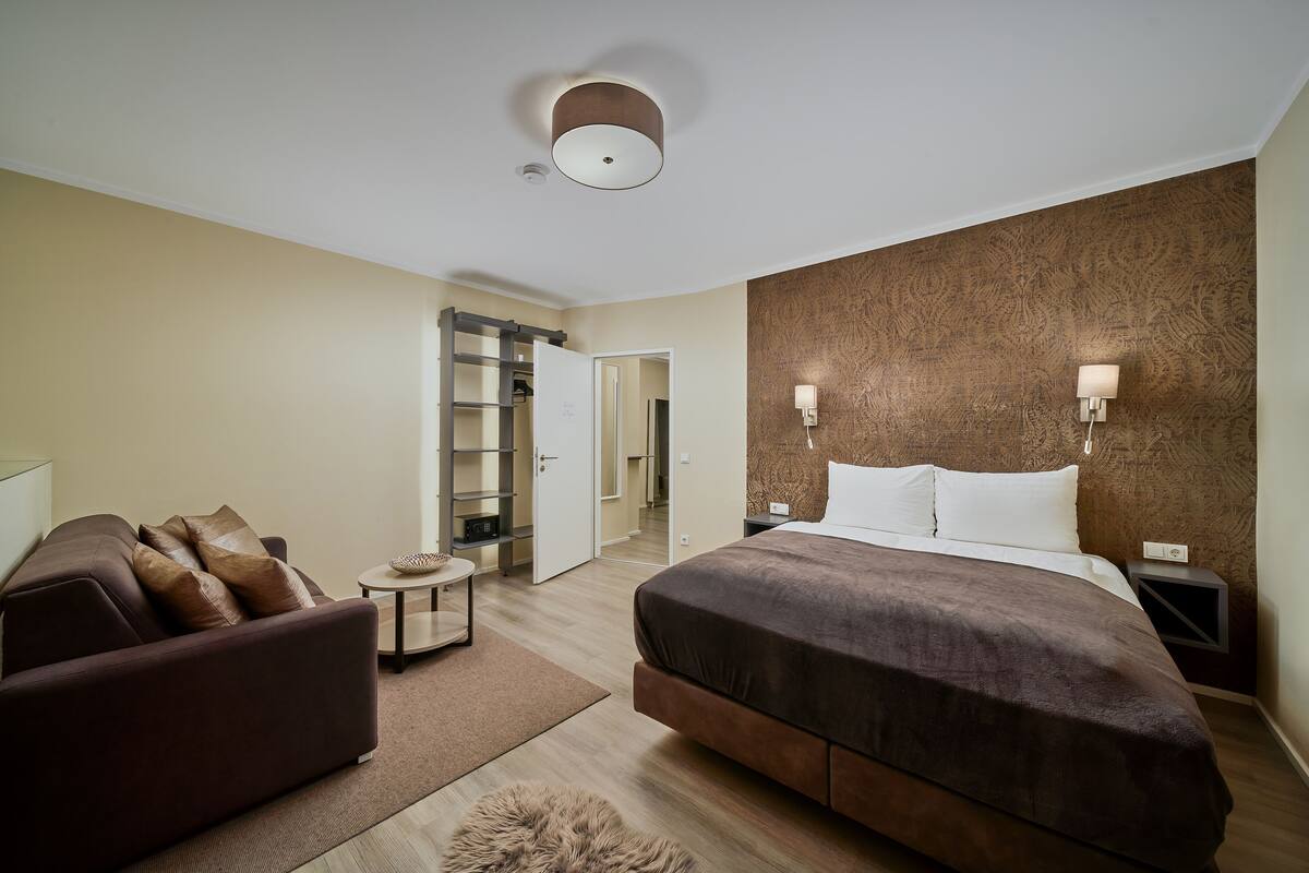 A modern hotel room with a large bed, a sofa, a small table, and a wardrobe.