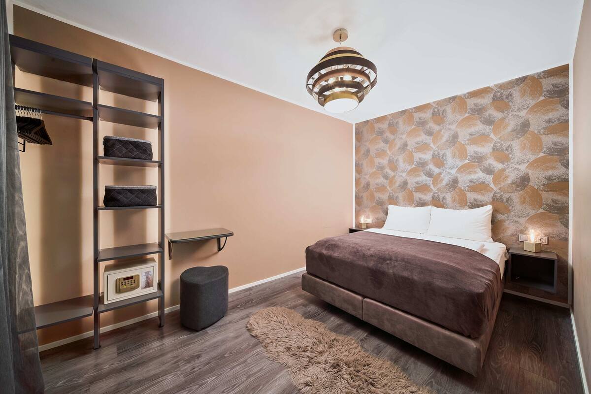 A bedroom with a bed, a wardrobe, a small table, and a wall-mounted shelf.