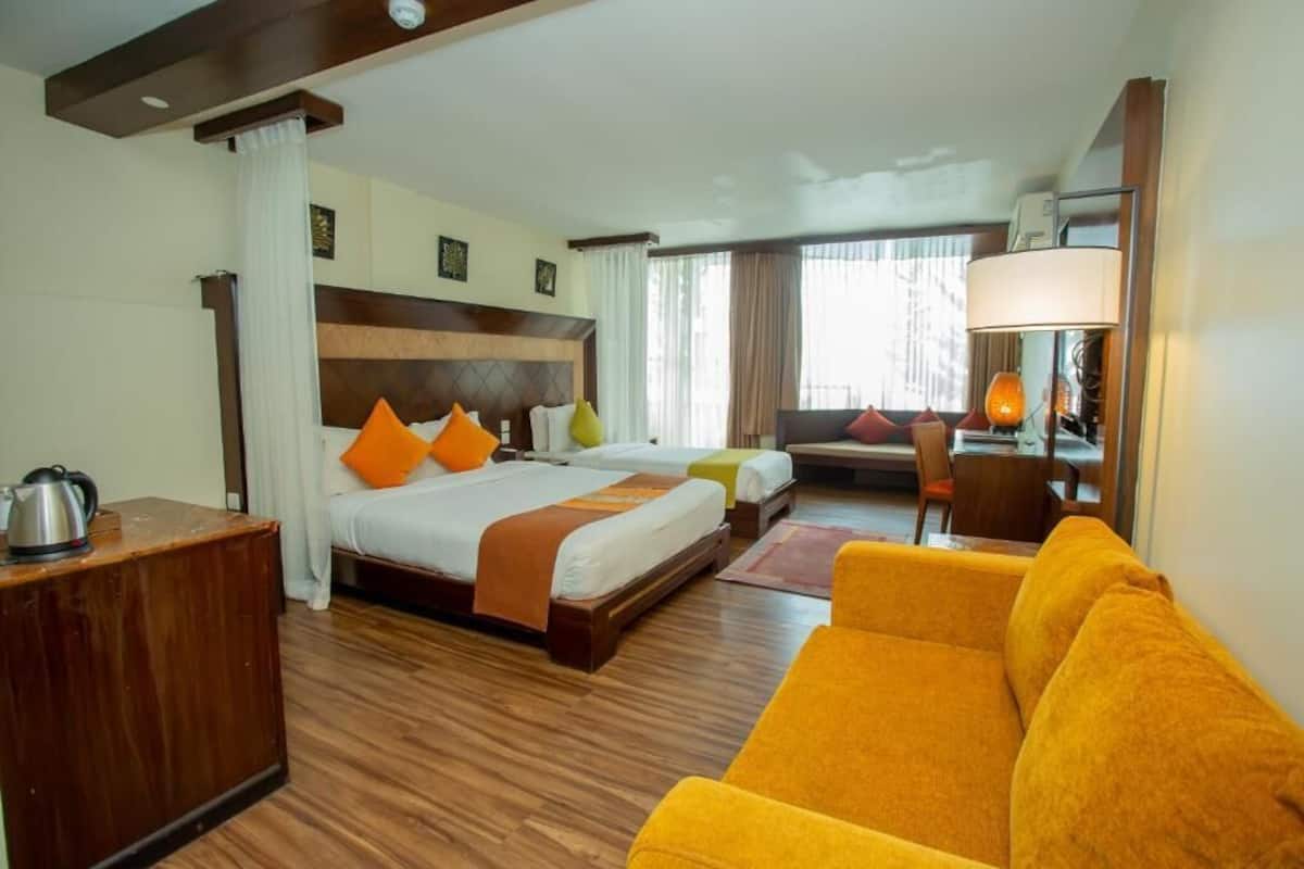 A hotel room with a wooden floor, a large bed with orange and white bedding, a yellow sofa, a wooden nightstand, and a lamp.