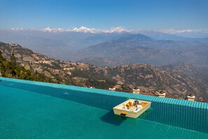 Una piscina infinita con una caja de madera que contiene bebidas y aperitivos, con vistas a un paisaje montañoso.