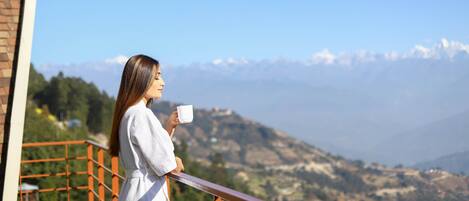 Una persona de pie en un balcón con barandilla, sosteniendo una taza, con vistas a un paisaje montañoso.