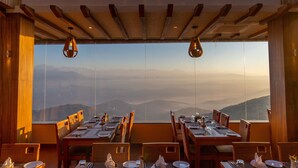 Un restaurante con vistas a montañas a través de amplios ventanales, vigas de techo de madera y sillas de mimbre.