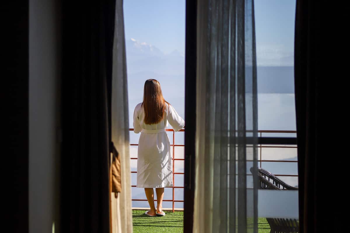 A person in a white dress standing at a window with a view of a body of water and mountains.