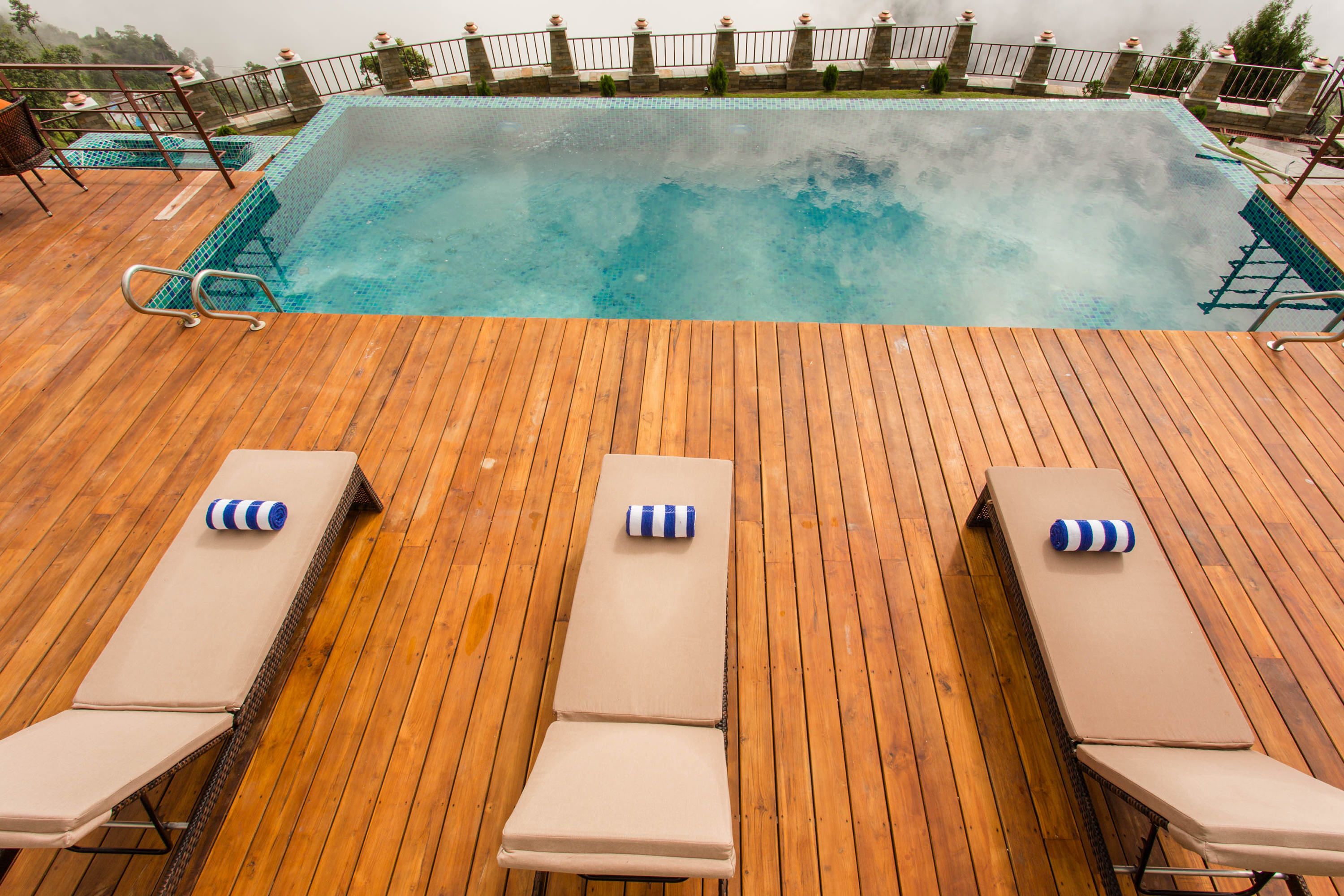 A wooden deck with a pool, lounge chairs, and a stone balustrade.