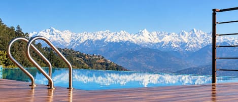 A wooden deck with a metal railing overlooking a clear blue lake and snow-capped mountains in the background.
