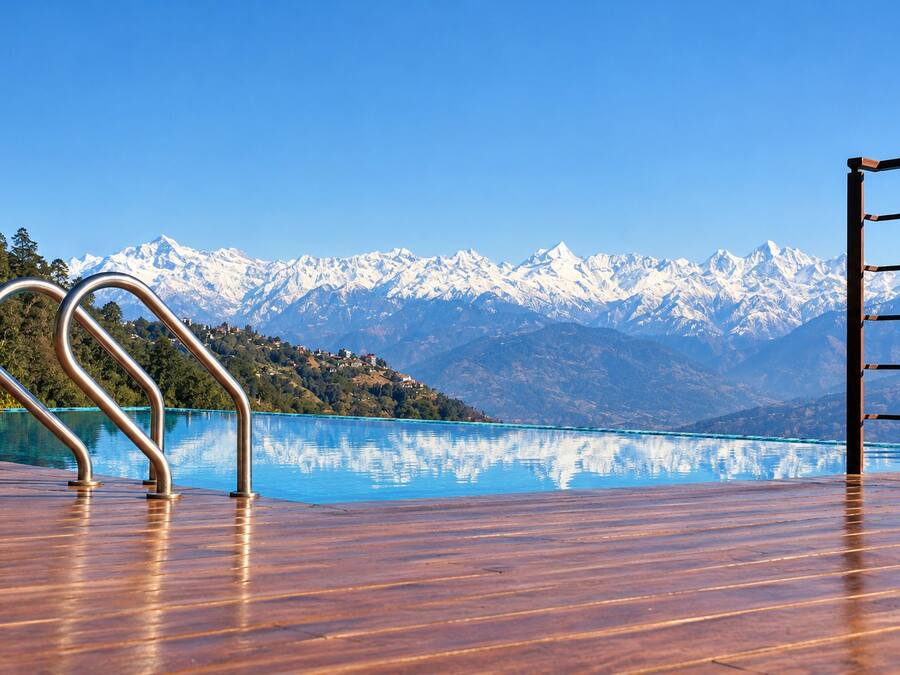A wooden deck with a metal railing overlooking a clear blue lake and snow-capped mountains in the background.