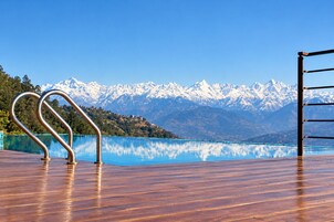 Um deck de madeira com guarda-corpo de metal, com vista para um lago azul límpido e montanhas cobertas de neve ao fundo.