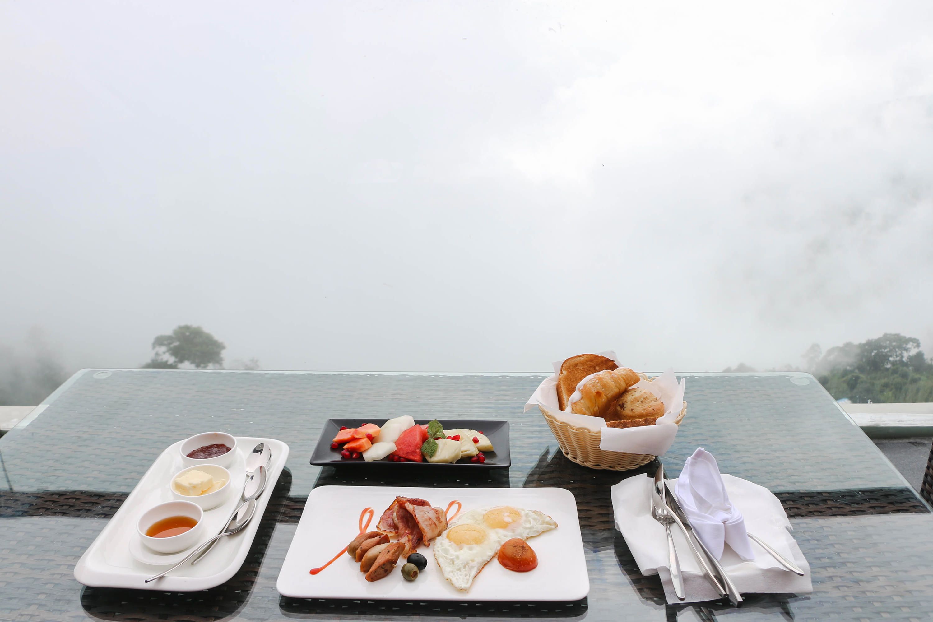 A breakfast table with plates of food, a basket of bread, and a view of the misty landscape.