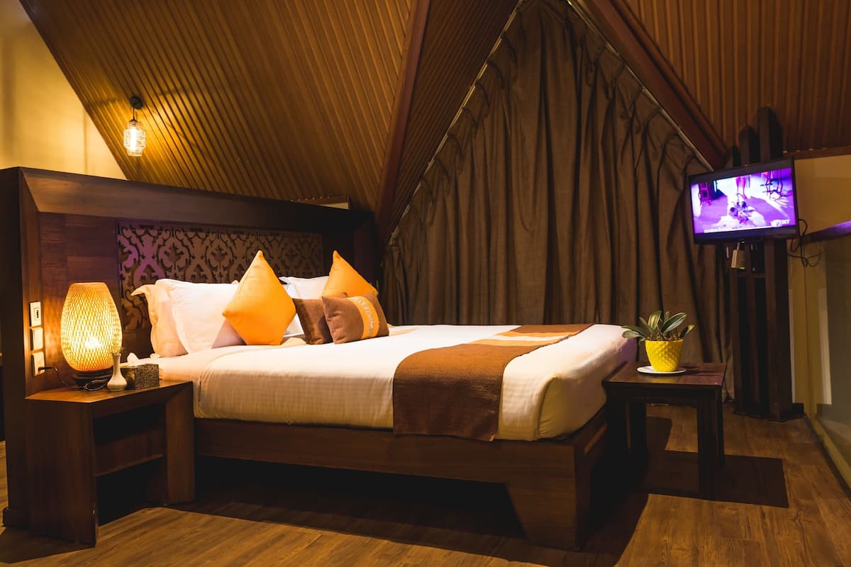 A bedroom with a wooden ceiling, a bed with white bedding and orange pillows, a bedside lamp, a TV on the right, and a small table with a plant.