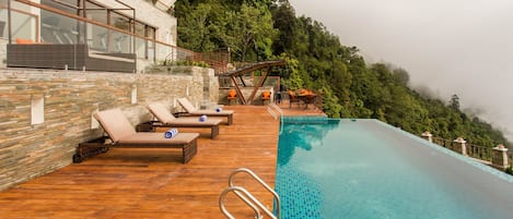 An infinity pool with a wooden deck and lounge chairs, surrounded by a stone wall and overlooking a forested hillside.