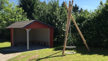 Un pequeño edificio blanco y rojo con un porche cubierto, un columpio y un trampolín circular en un césped.