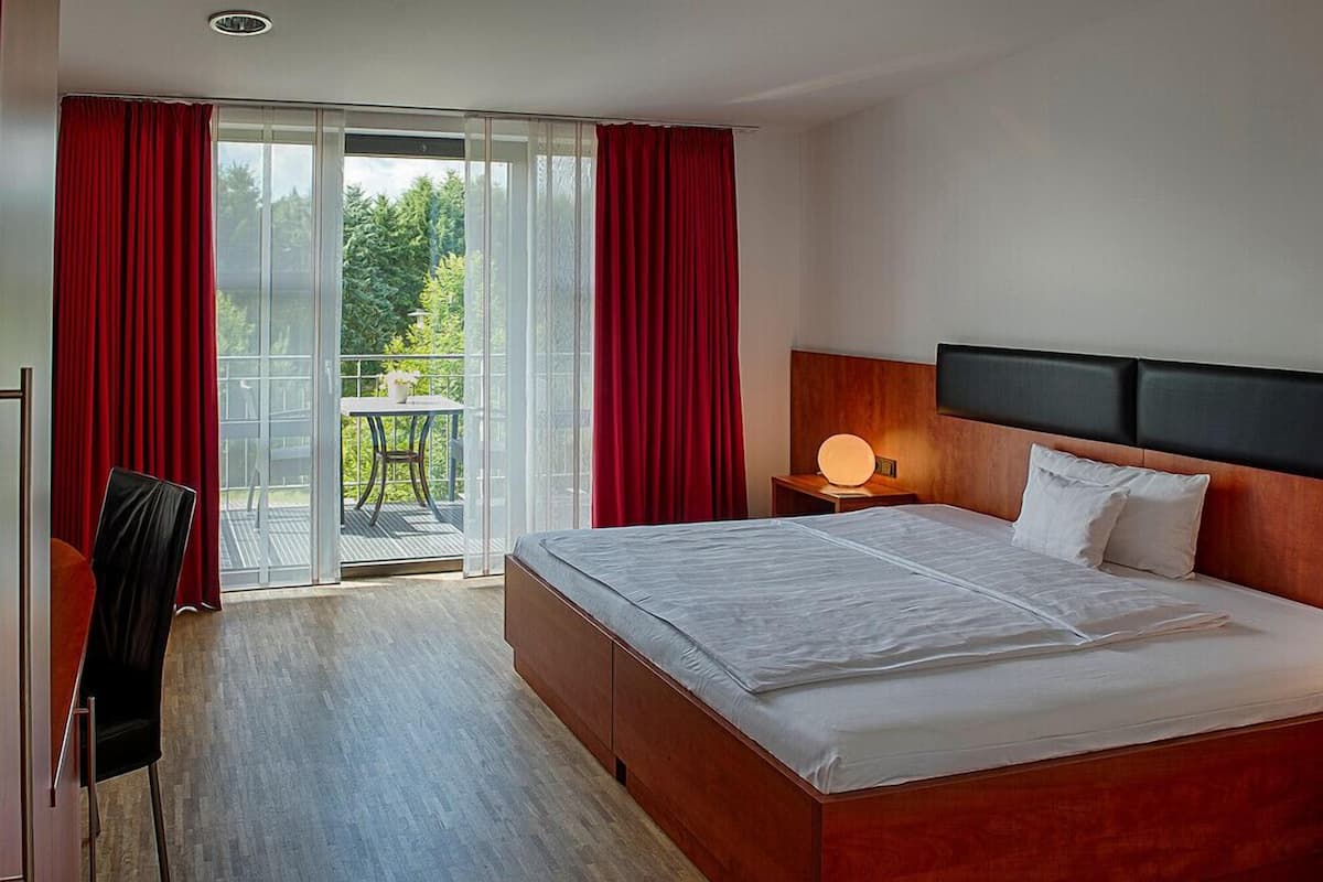 A hotel room with a large bed, a nightstand with a lamp, a chair, and a balcony with red curtains.