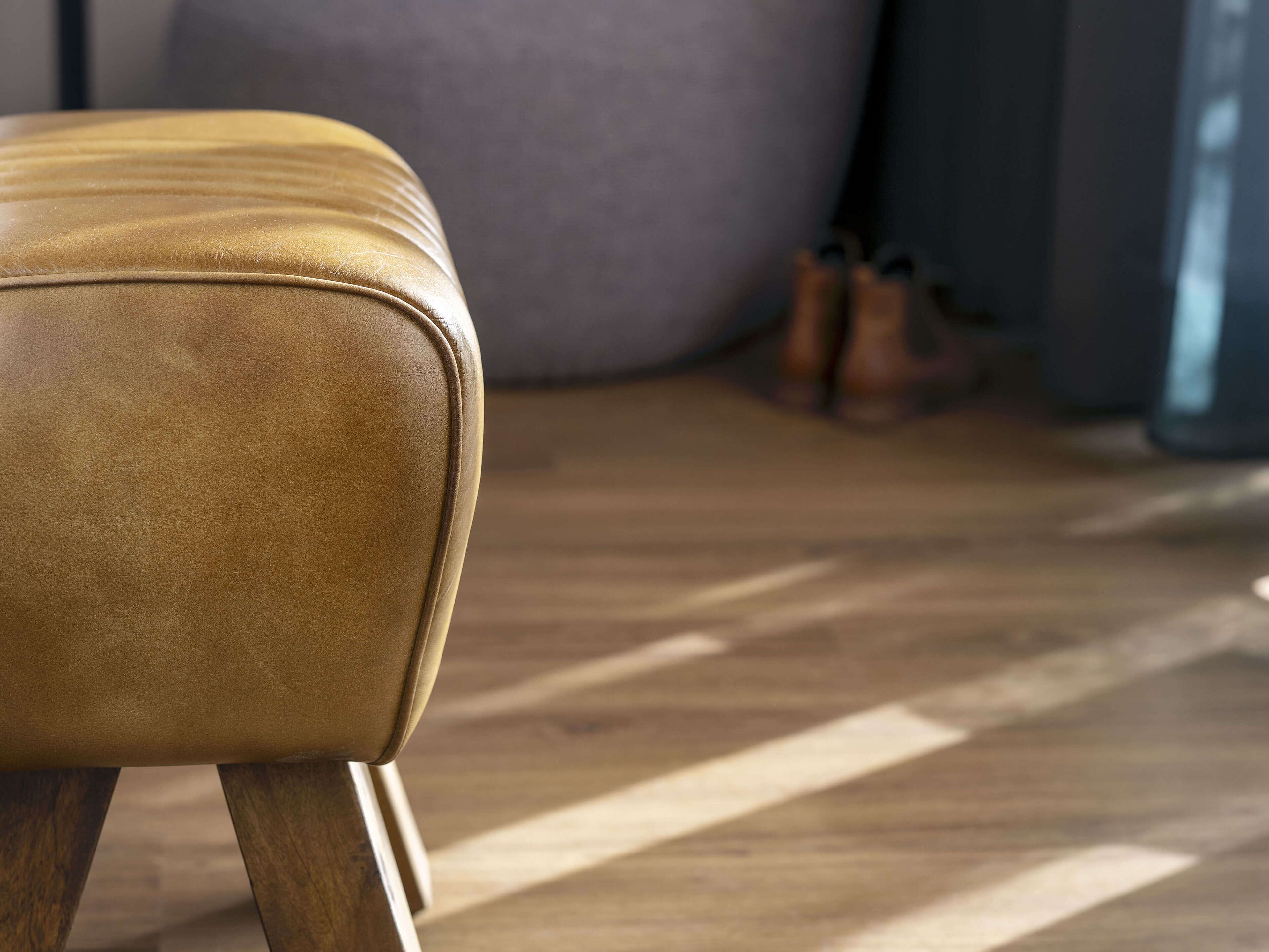 Un canapé en cuir avec des pieds en bois, dans une pièce dont le sol est également en bois, et un grand coussin gris en arrière-plan.