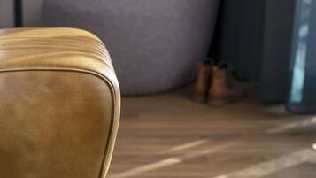 Un canapé en cuir avec des pieds en bois, dans une pièce dont le sol est également en bois, et un grand coussin gris en arrière-plan.
