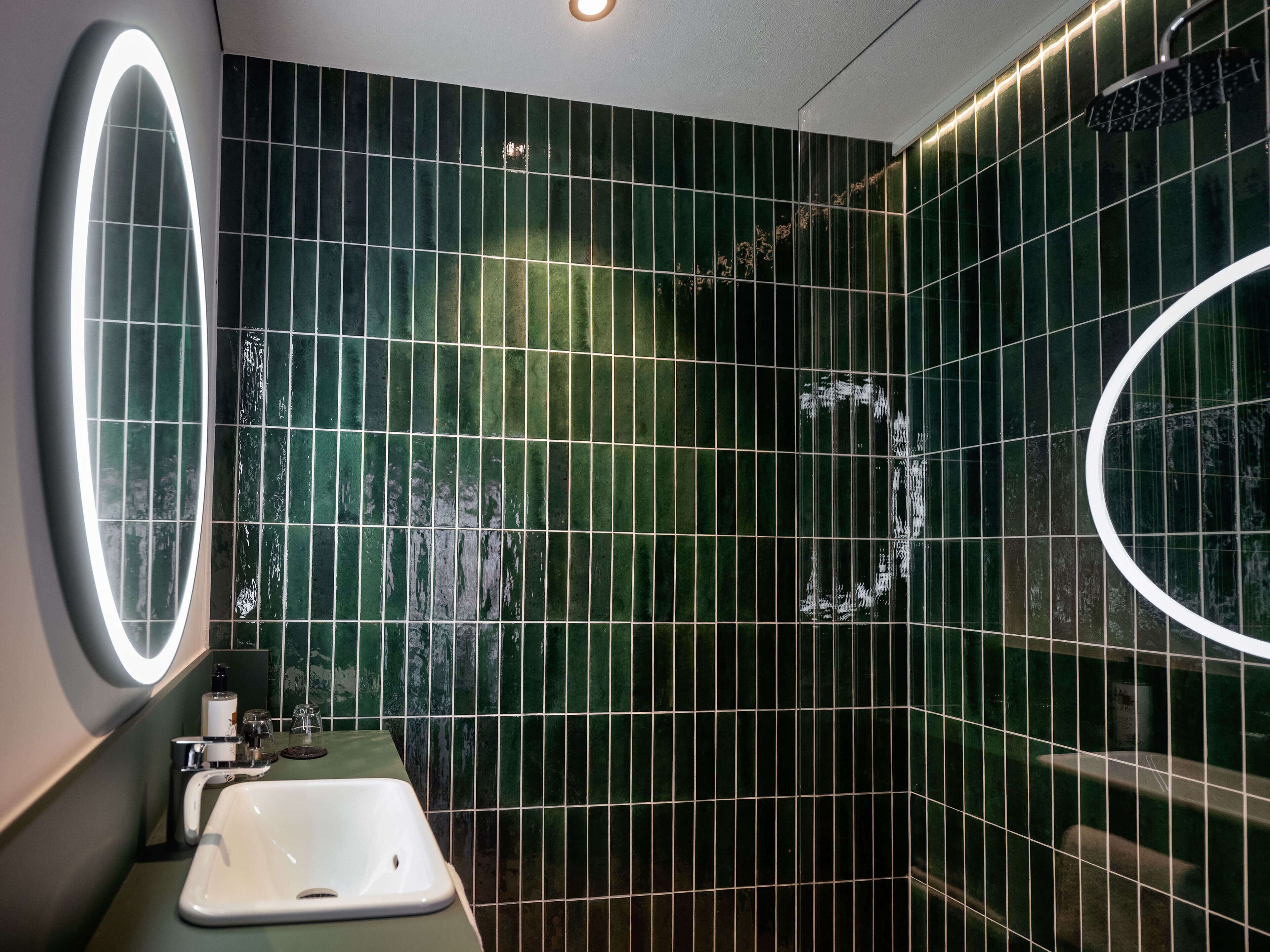 Un baño con baldosas verde oscuro, un lavamanos blanco y un espejo con luz circular.