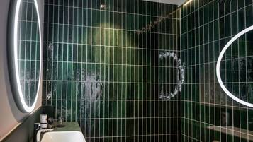 Un baño con baldosas verde oscuro, un lavamanos blanco y un espejo con luz circular.