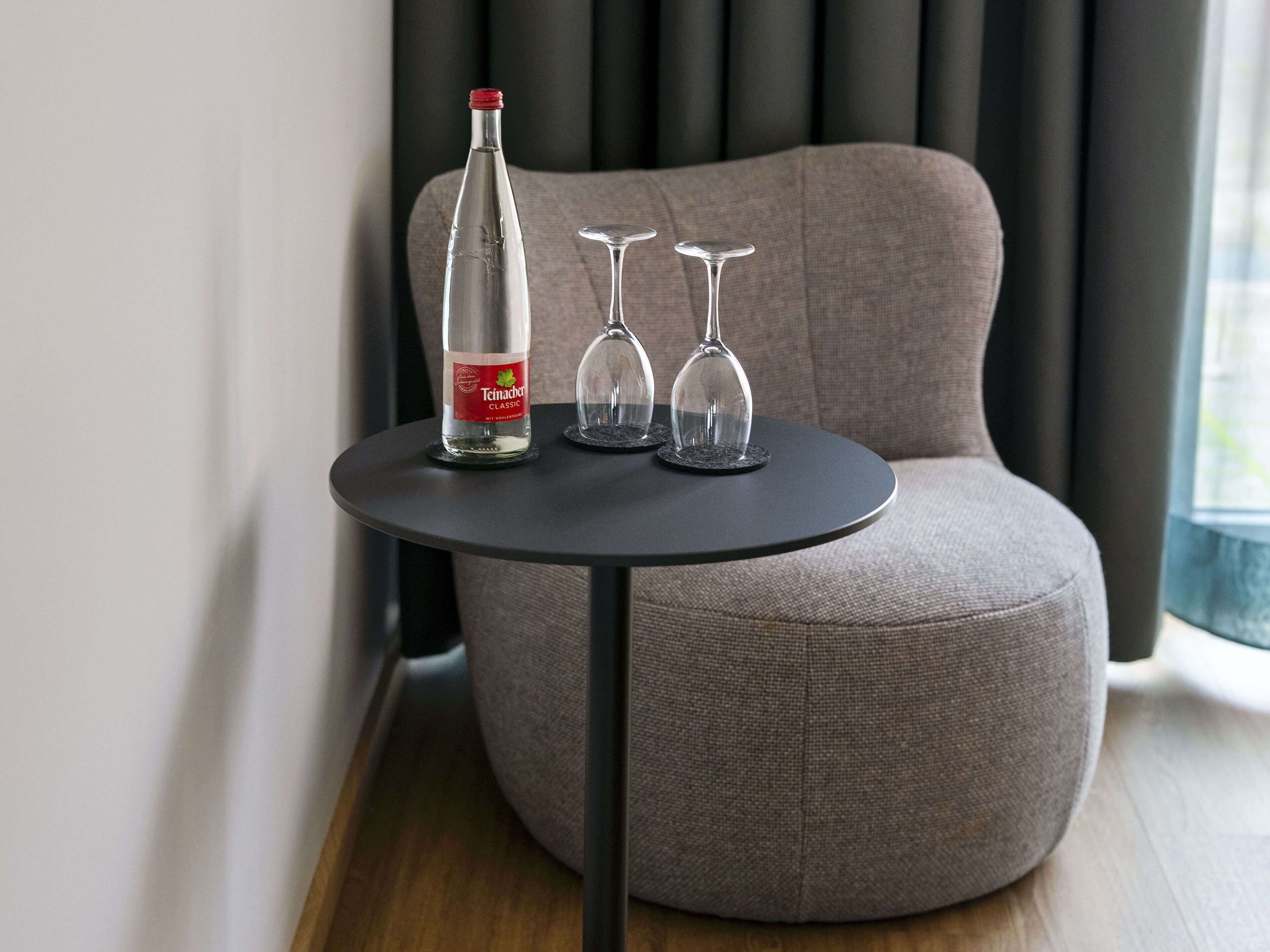 Una habitación moderna con un sofá redondo gris, una mesita negra, una botella de vino Teimacher Classic y dos copas de vino.