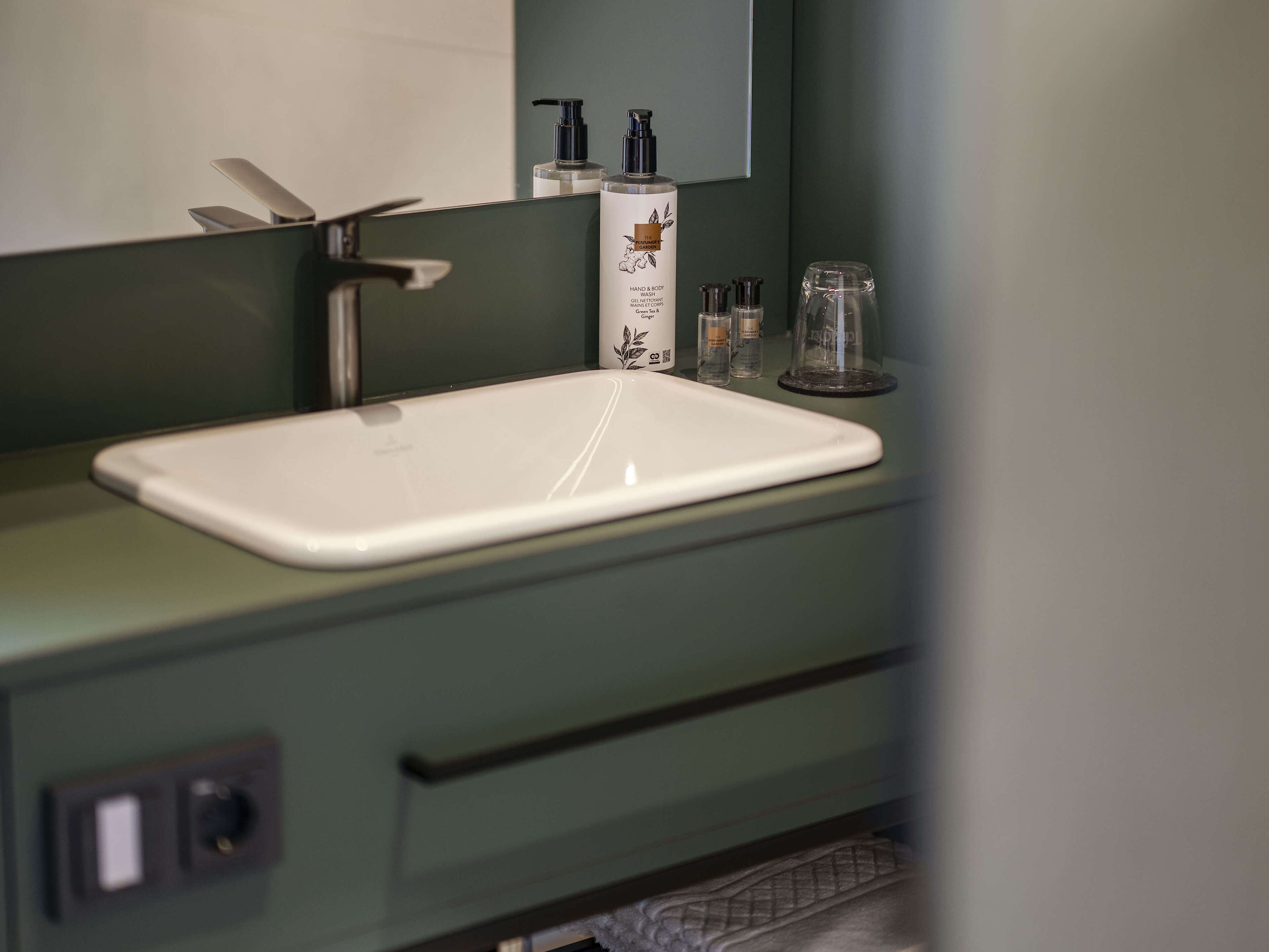 Un baño con un lavamanos blanco, encimera verde y un espejo arriba. Hay artículos de aseo en la encimera.