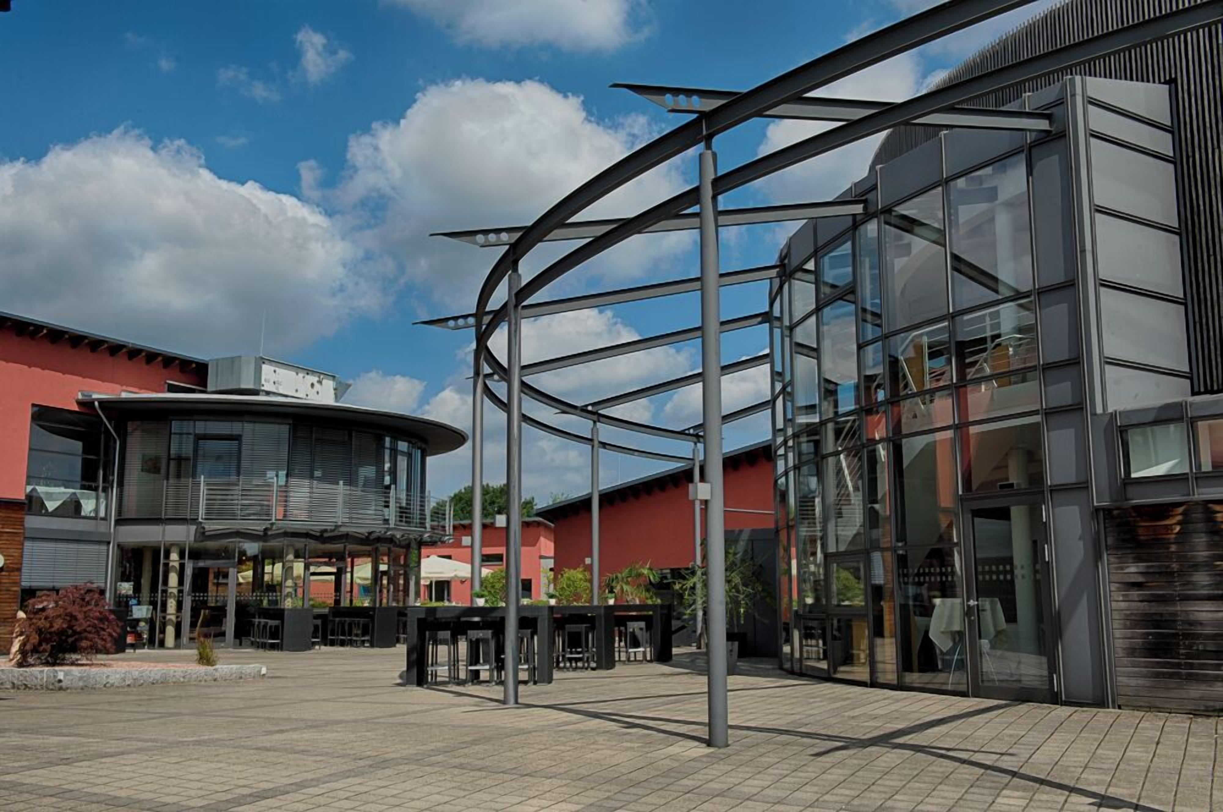 A modern building with a glass facade, a curved metal structure, and a red building in the background.