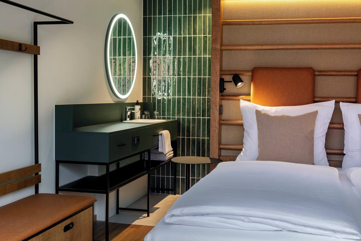 A modern hotel room with a large bed, a vanity with a mirror, a small round table, and a bench.