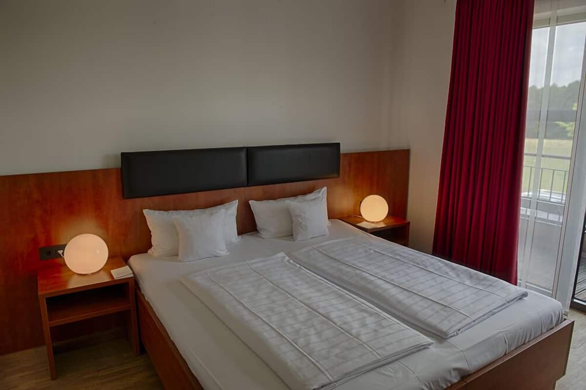 A hotel room with a large bed, two bedside tables with lamps, a wooden headboard, and a view of the outdoors through a sliding glass door.